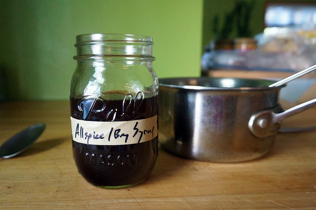 The deep, brooding heart of the drink: a jar labeled 'Allspice/Bay Syrup' is half-filled with an opaque brown liquid, in front of a saucepan with a ladle resting in it.