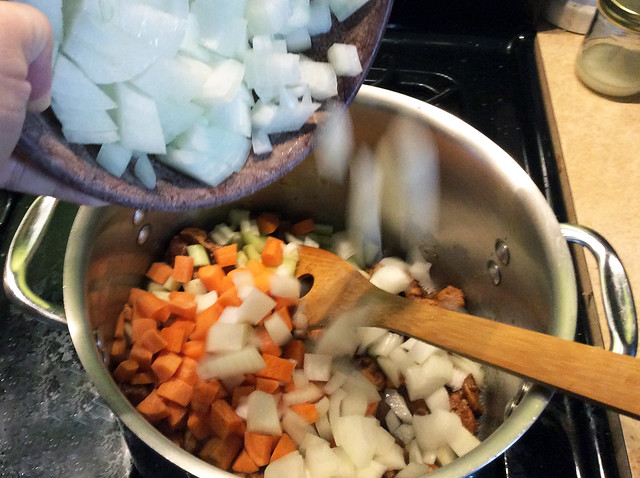 A tumble of white onions cascades from a prep bowl into a large pot, the falling ones a blur.