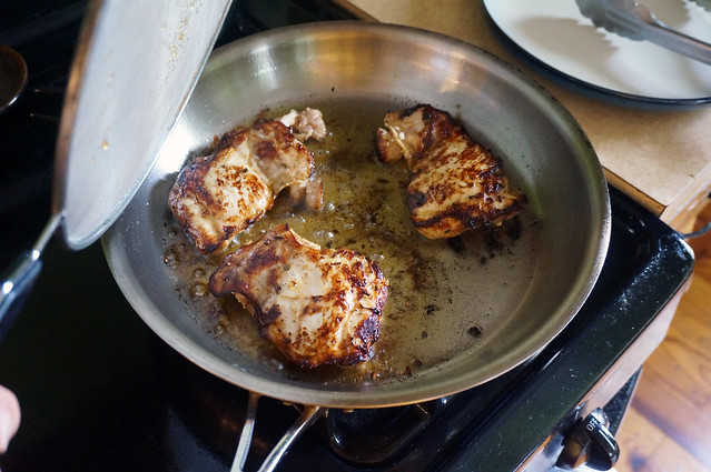 A large stainless steel skillet sits on a black enamel stovetop, with three chicken thighs bubbling away in the olive oil.