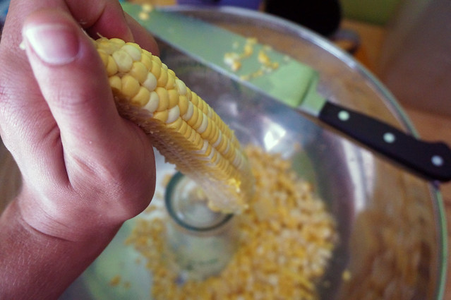 Shot from above, my kernel-stripping setup: a very large bowl with a pint glass upside down in the bottom. My hand holds up a corncob, half-stripped, resting on the edge of the pint glass, the knife waiting to slice off the rest of the kernels