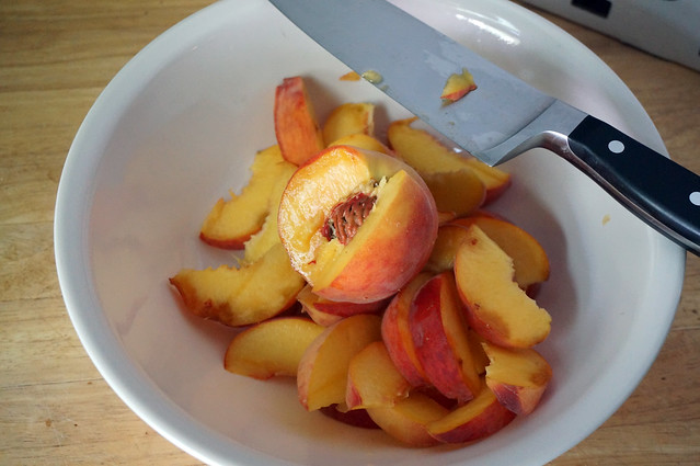 A wide, white bowl half-filled with peach slices. A partially sliced peach, russet pit exposed, sits on top of the pile, while a large chef's knife rests delicately across the rim. This bowl fills me with childlike glee.